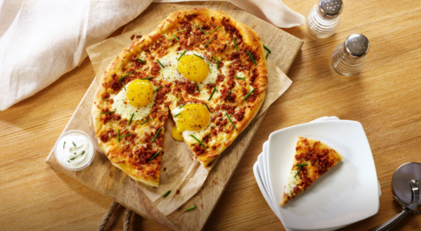 oval shaped pizza with a slice removed to the sides with three sunny side up eggs on top and a small sour cream with chives sauce next to the pizza