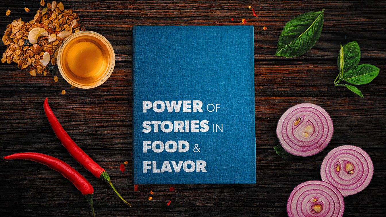 Blue hard bound book with Power of Stories in Food & Flavor text placed in the center of a dark wooden table with two chili peppers, granola and honey in a sauce cup, two fresh basils and three sliced red onions