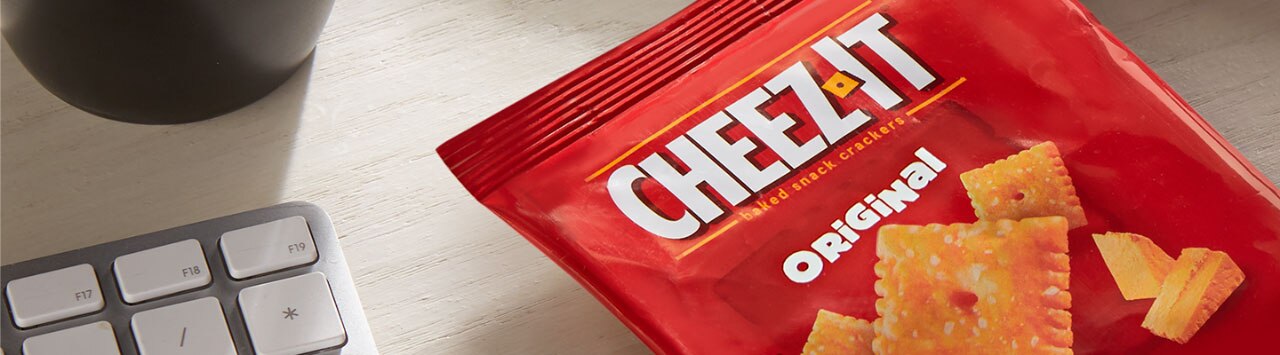 A small Cheez-It original flavor pouch placed diagonally on the bottom right next to a white and gray keyboard and a bottom black mug